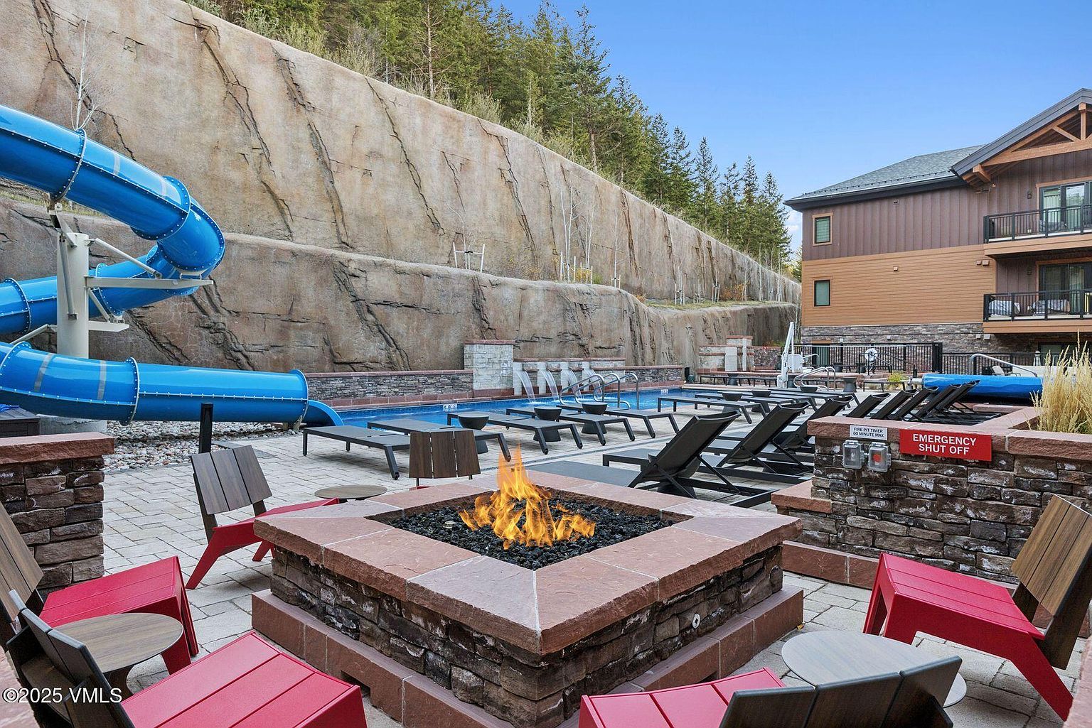 This image showcases a luxurious outdoor pool and spa area, complete with a water slide and a cozy fire pit surrounded by comfortable seating. The pool is lined with lounge chairs, and the area is nestled against a natural rock wall with trees, creating a serene and private atmosphere. The building in the background suggests a resort or high-end residential complex.