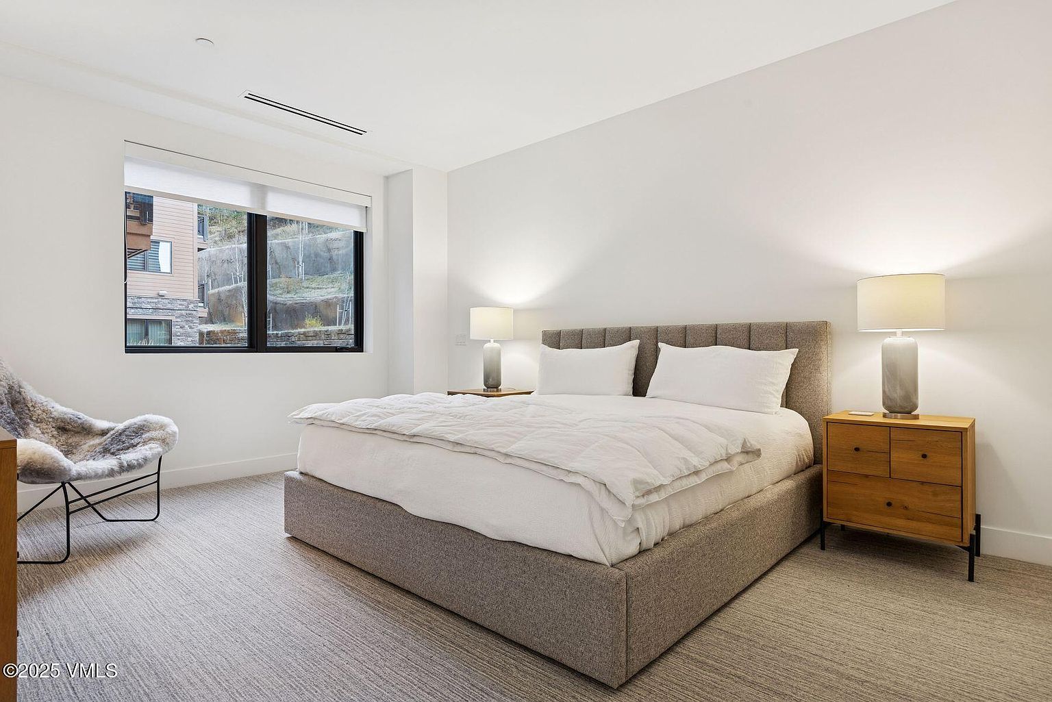 This is a bright and airy bedroom featuring a queen-sized bed with a gray upholstered frame and headboard, crisp white linens, and two matching nightstands with lamps. A modern chair sits near the window, which provides natural light. The room has a clean, minimalist aesthetic with neutral tones and carpet flooring.