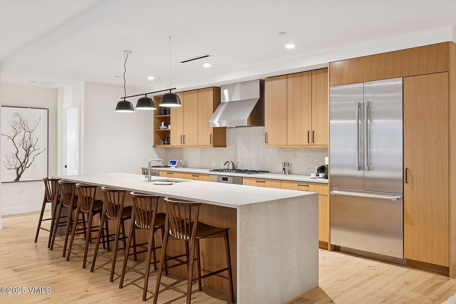 This is a bright and modern kitchen featuring light wood cabinetry, stainless steel appliances, and a large island with a light-colored countertop. The kitchen is well-lit with pendant lighting above the island and recessed lighting throughout. The overall impression is clean, spacious, and inviting, perfect for cooking and entertaining.