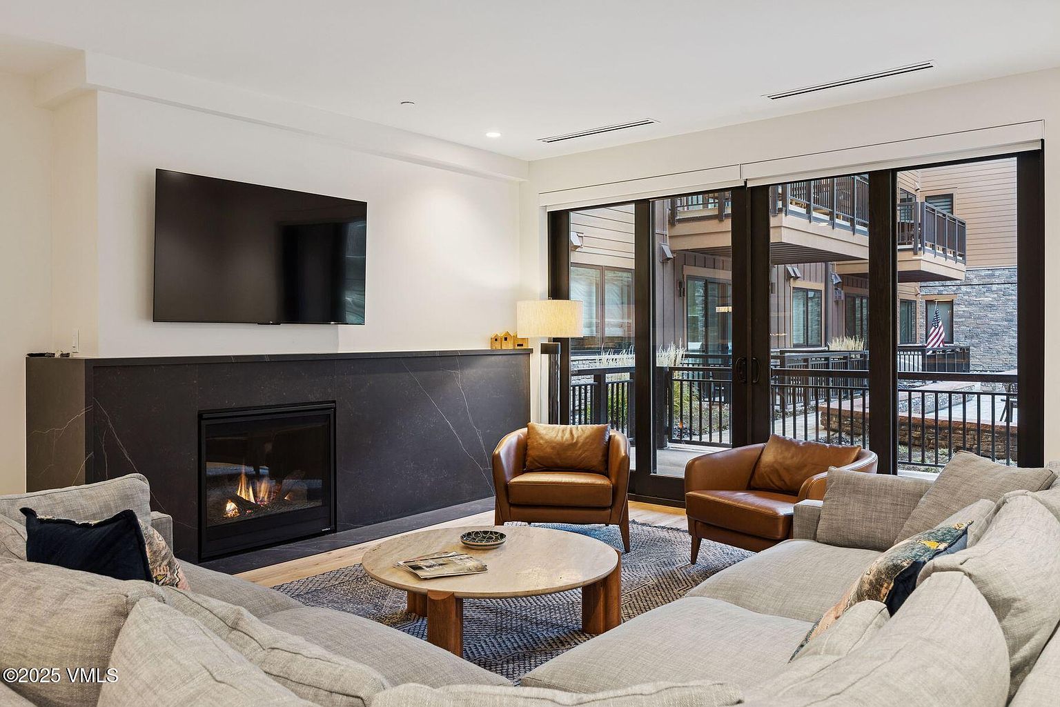This is an interior shot of a living room featuring a modern fireplace with a large flat-screen TV mounted above it. The room is furnished with a comfortable sectional sofa, two leather armchairs, and a round coffee table on a patterned rug. Large sliding glass doors provide natural light and a view of the outdoor balcony and surrounding buildings, creating a cozy and inviting atmosphere.