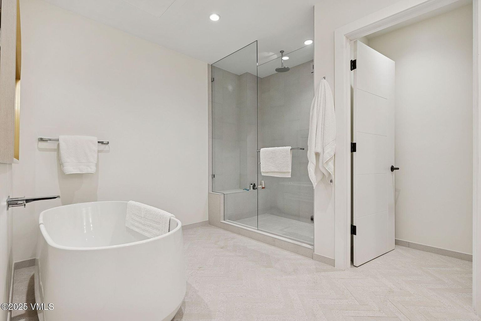 This is a bright and modern primary bathroom featuring a freestanding white bathtub, a glass-enclosed shower with a rainfall showerhead, and neutral-toned herringbone tile flooring. The walls are painted in a soft white, enhancing the sense of spaciousness and cleanliness. The door is slightly ajar, inviting the viewer into the space.