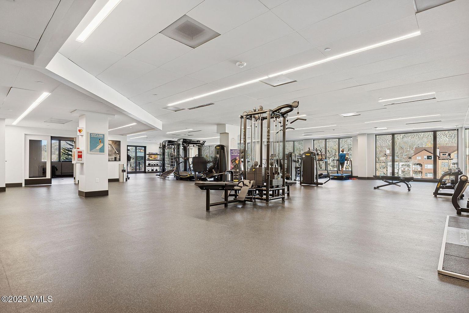 This is a spacious home gym featuring a variety of exercise equipment, including weight machines, treadmills, and benches. The room has a neutral color palette with a gray floor and white ceiling, and large windows provide natural light. The overall impression is clean, well-equipped, and conducive to a workout.
