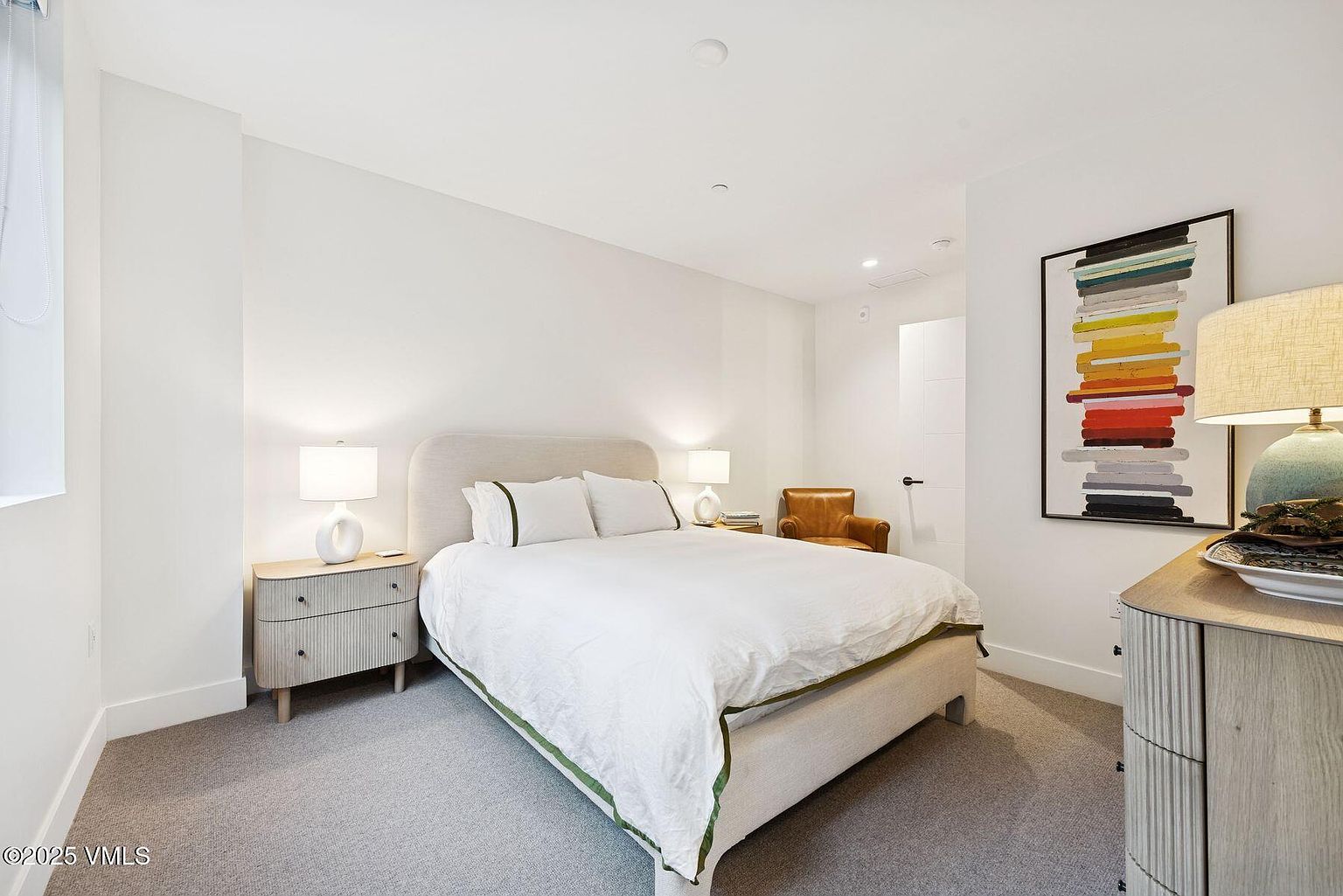 This is a bright and airy primary bedroom featuring a neutral color palette. The room includes a bed with white linens and green trim, two nightstands with lamps, and a modern art piece on the wall. A tan leather chair is visible in the background, adding a touch of warmth to the space.