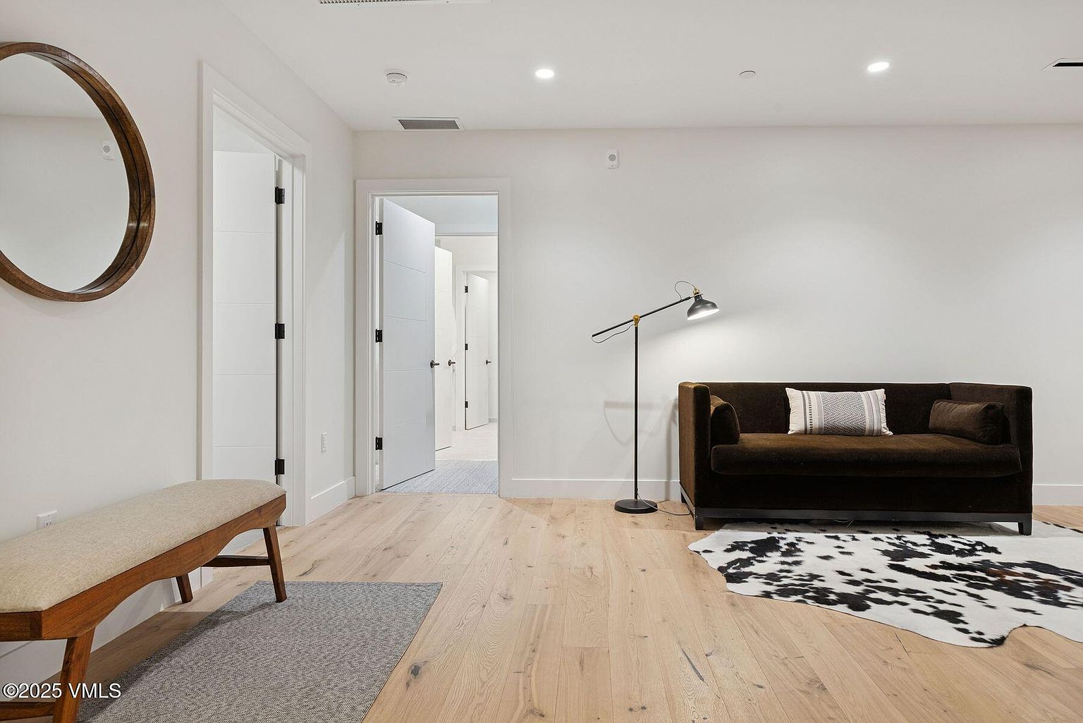 This interior shot showcases a modern living room with light hardwood floors and white walls. A dark brown sofa is positioned against the wall, complemented by a floor lamp and a black and white cowhide rug. A wooden bench and a round mirror add to the room's aesthetic, creating a stylish and inviting space.