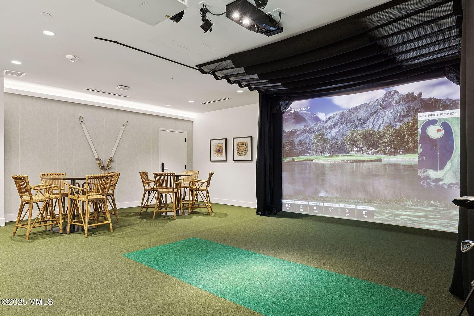 This interior shot showcases a home golf simulator room, featuring a large projection screen displaying a golf course scene. The room includes seating areas with wicker chairs and tables, creating a comfortable space for relaxation and entertainment. The green carpet and putting area enhance the golfing experience, while the overall design is modern and inviting.
