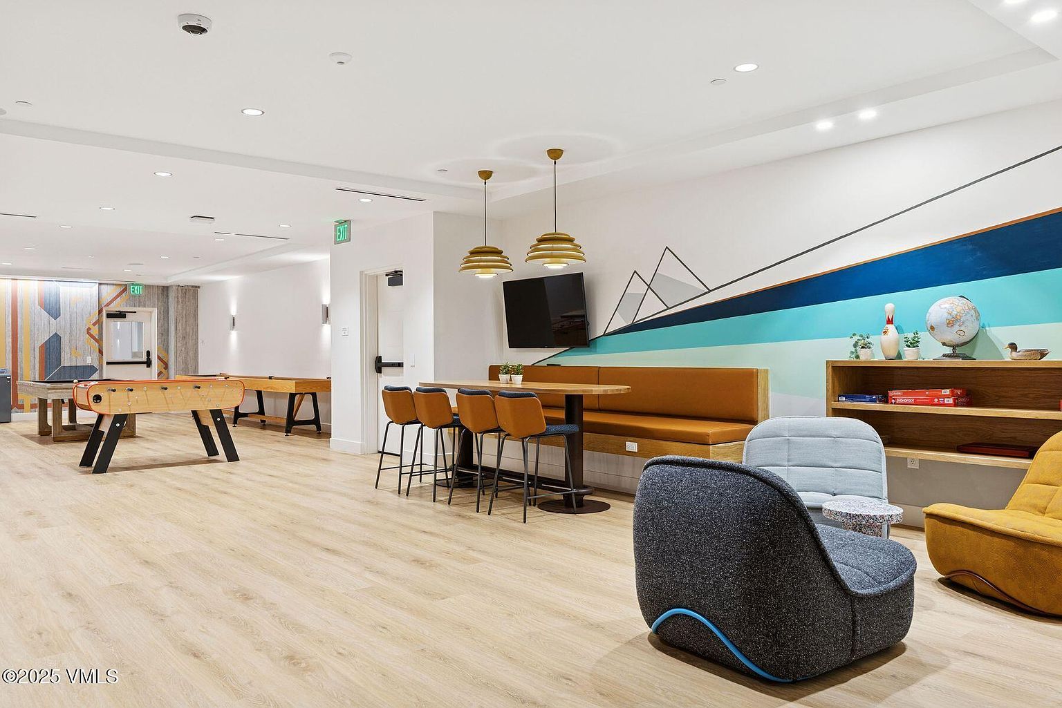 This is a well-lit community amenity space featuring a foosball table, a dining area with bar stools and a booth, and comfortable lounge seating. The room is decorated with a modern mountain mural and includes a bookshelf with decorative items, creating a welcoming and recreational environment. The light wood flooring and ample lighting contribute to the bright and airy atmosphere.