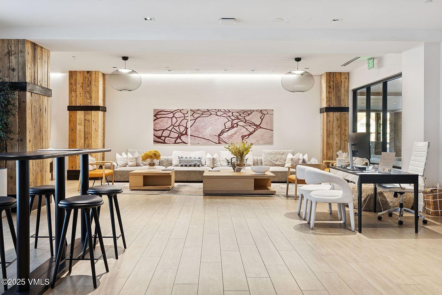 This is a well-lit living room featuring a neutral color palette with wooden accents. The room includes multiple sofas, coffee tables, and decorative artwork, creating a comfortable and inviting atmosphere. The flooring is light-colored wood, and the space is illuminated by modern pendant lights.