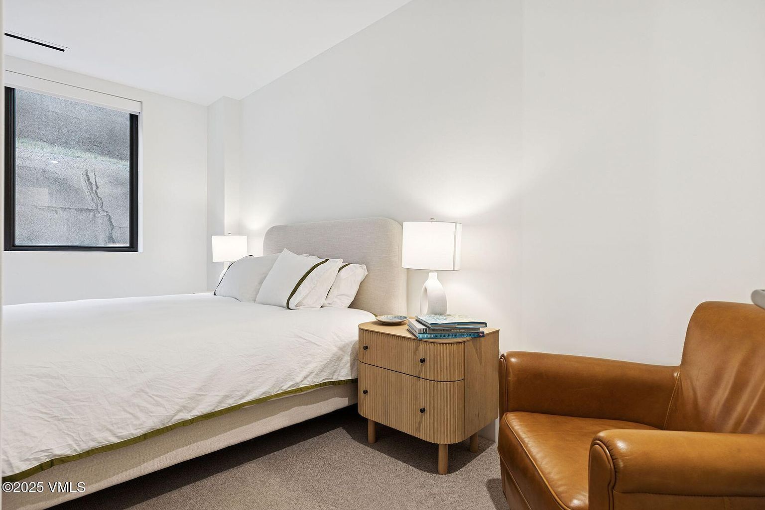 This is a bright and minimalist guest bedroom featuring a bed with white linens and a light gray headboard. A modern nightstand with a lamp sits beside the bed, and a tan leather armchair is positioned in the corner. The room has a clean and uncluttered feel, with a window providing natural light.