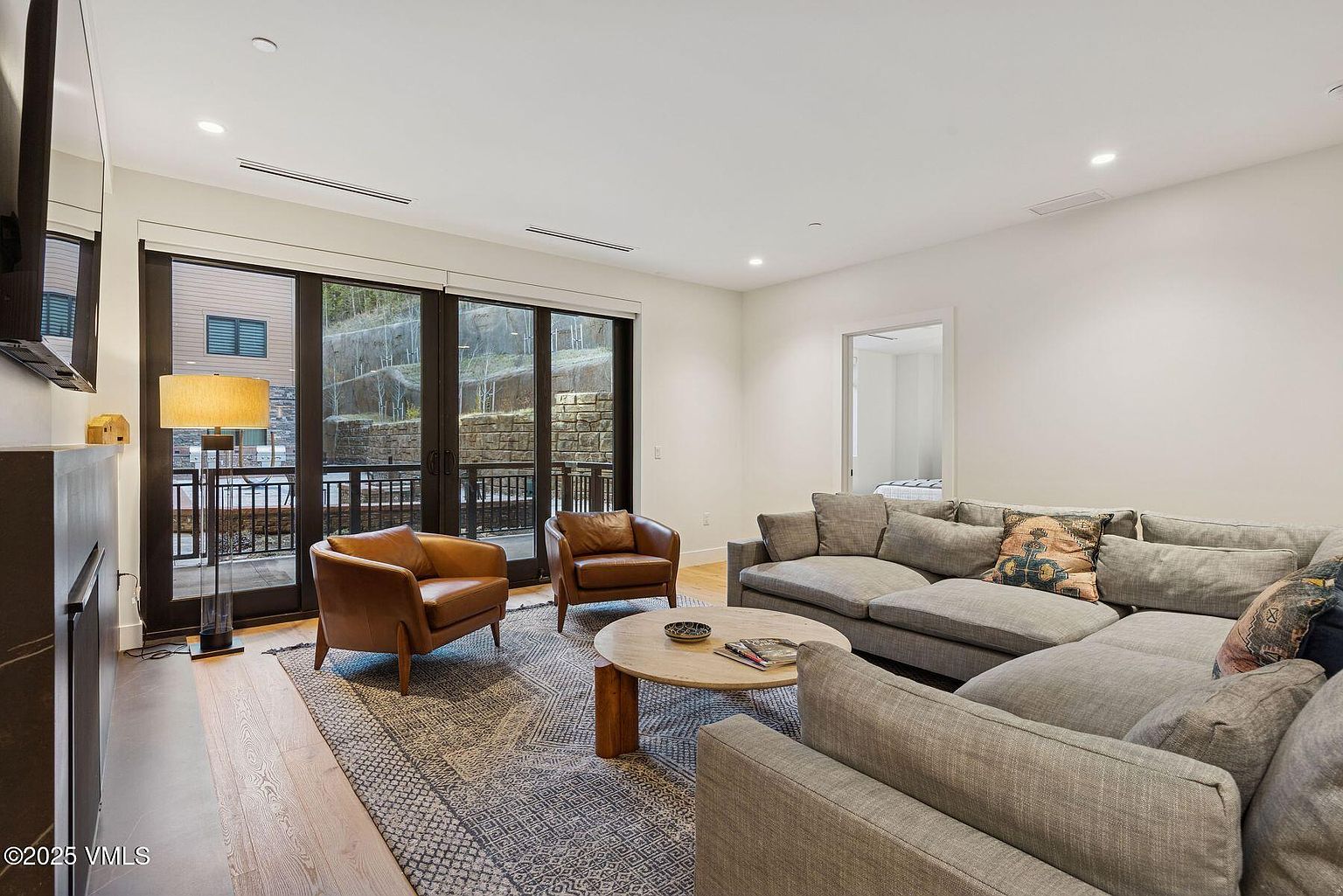 This is an interior shot of a living room featuring a large gray sectional sofa, two leather armchairs, and a round wooden coffee table. The room has a modern aesthetic with a neutral color palette, hardwood floors, and a large sliding glass door leading to an outdoor area. The space is well-lit with recessed lighting and a floor lamp, creating a warm and inviting atmosphere.