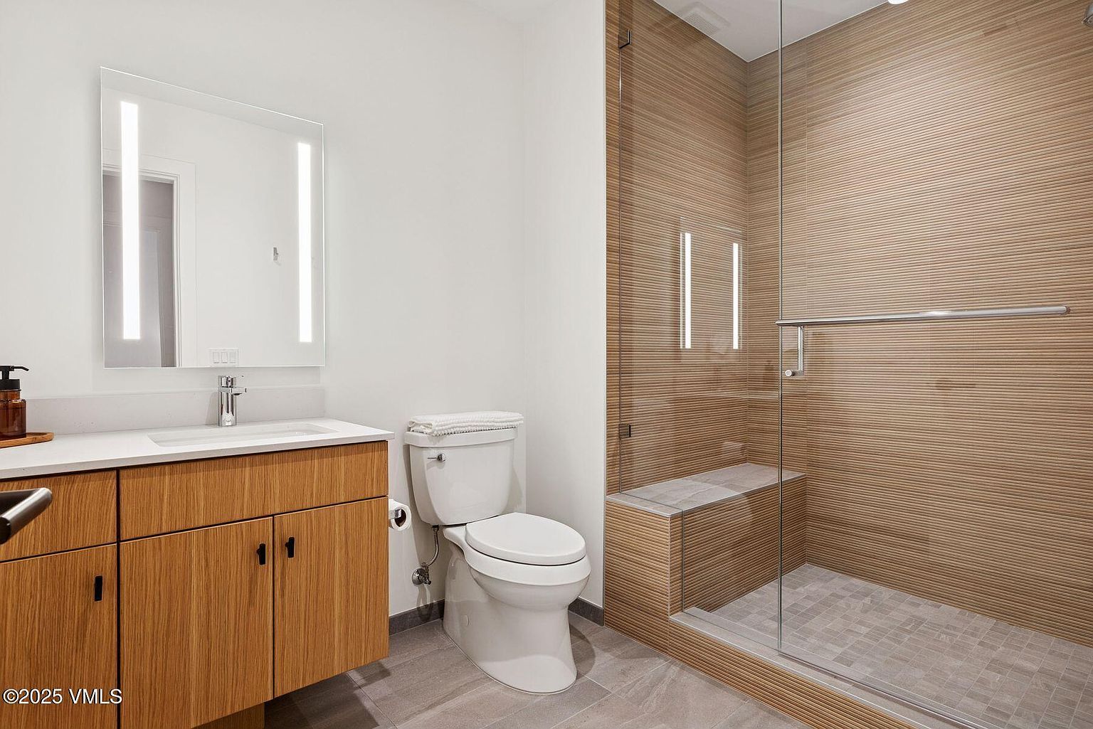 This is a modern bathroom featuring a floating wood vanity with a white countertop and a large, illuminated mirror. A white toilet sits next to the vanity, and a glass-enclosed shower with horizontally-patterned wood-look tile and a built-in bench is visible. The floor is tiled in a neutral gray, creating a clean and contemporary aesthetic.