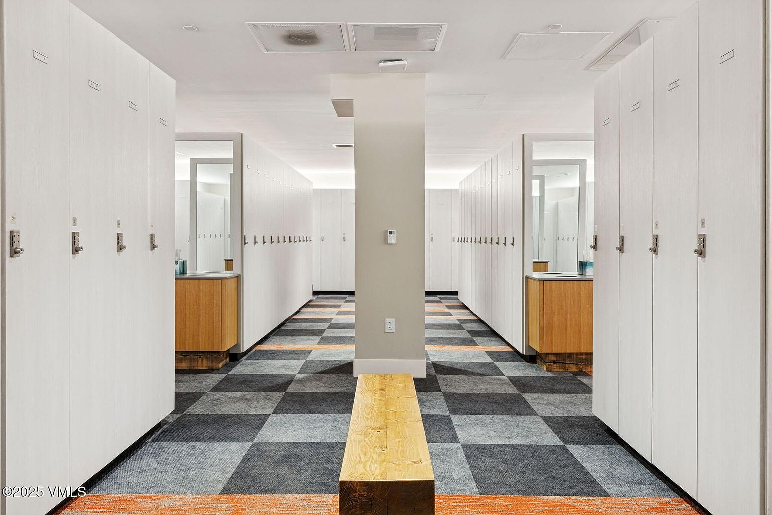 This is an interior shot of a locker room hallway, featuring rows of white lockers on either side. The floor is covered in a checkered pattern of gray and black tiles with orange accents along the edges. A wooden bench sits in the center of the hallway, and a support column is visible in the middle distance, adding to the symmetry of the space.