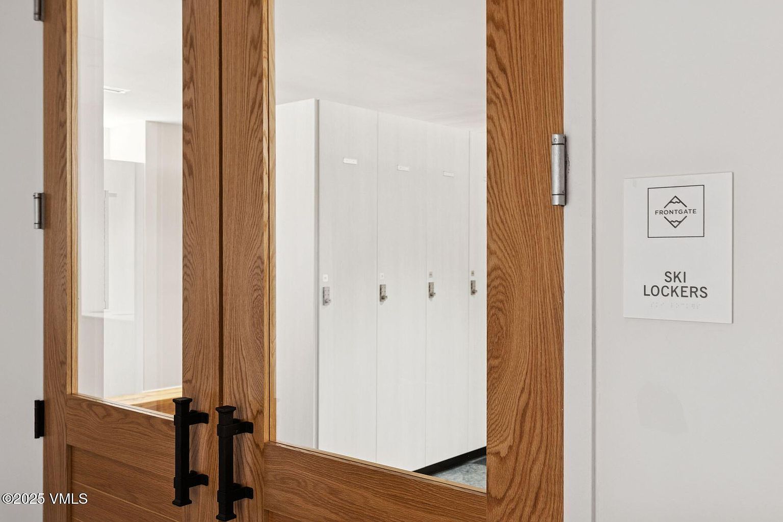 The image showcases a hallway area with a focus on ski lockers. The lockers are white and line the wall, visible through a wooden-framed glass door. A sign indicating "SKI LOCKERS" is mounted on the wall, adding to the functionality of the space.