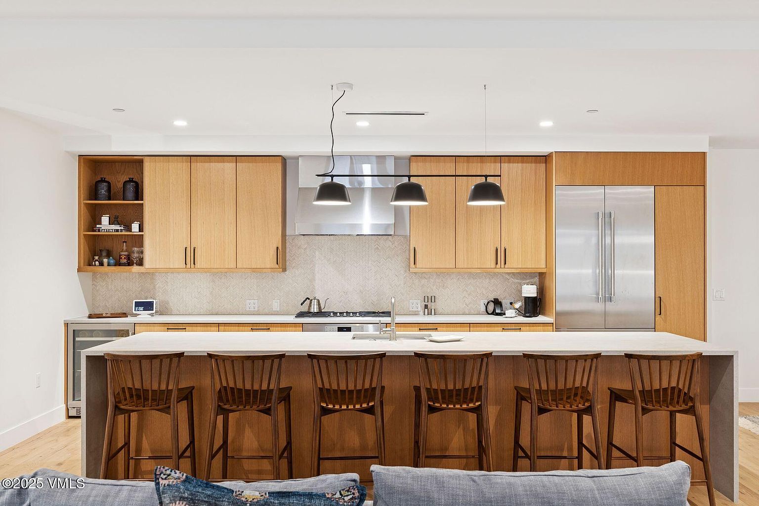 This is a well-lit, modern kitchen featuring light wood cabinetry and a large island with seating for six. Stainless steel appliances, including a refrigerator and range hood, add a sleek touch. The kitchen is designed with a clean aesthetic, and the bar stools provide a welcoming space for dining or socializing.