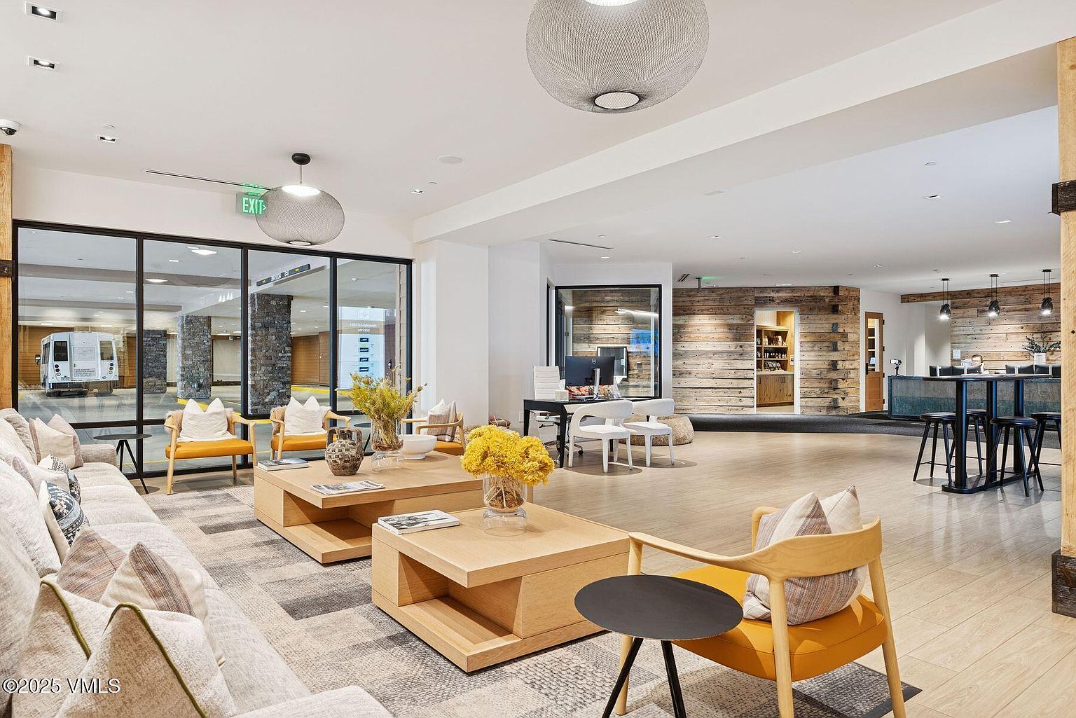 This is a well-lit, modern living room featuring a comfortable sofa with decorative pillows, wooden-framed chairs with yellow cushions, and stylish wooden coffee tables. Large windows offer an outside view, and contemporary light fixtures hang from the ceiling. The space is inviting and designed for relaxation and socializing.