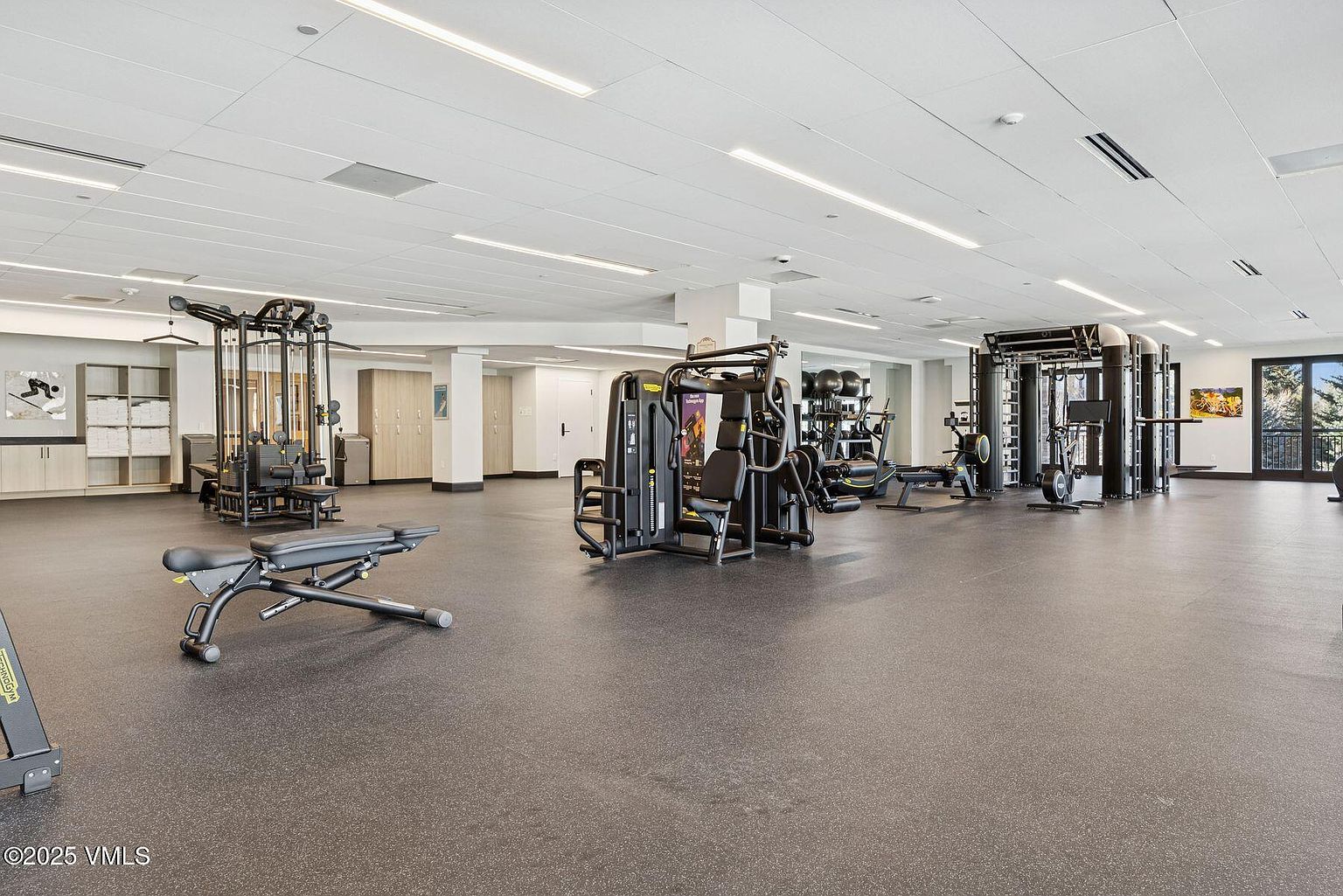This is a spacious home gym featuring a variety of exercise equipment, including weight machines, benches, and free weights. The flooring is a dark, durable material, and the lighting is bright and even. The overall impression is a well-equipped and functional space for fitness activities.