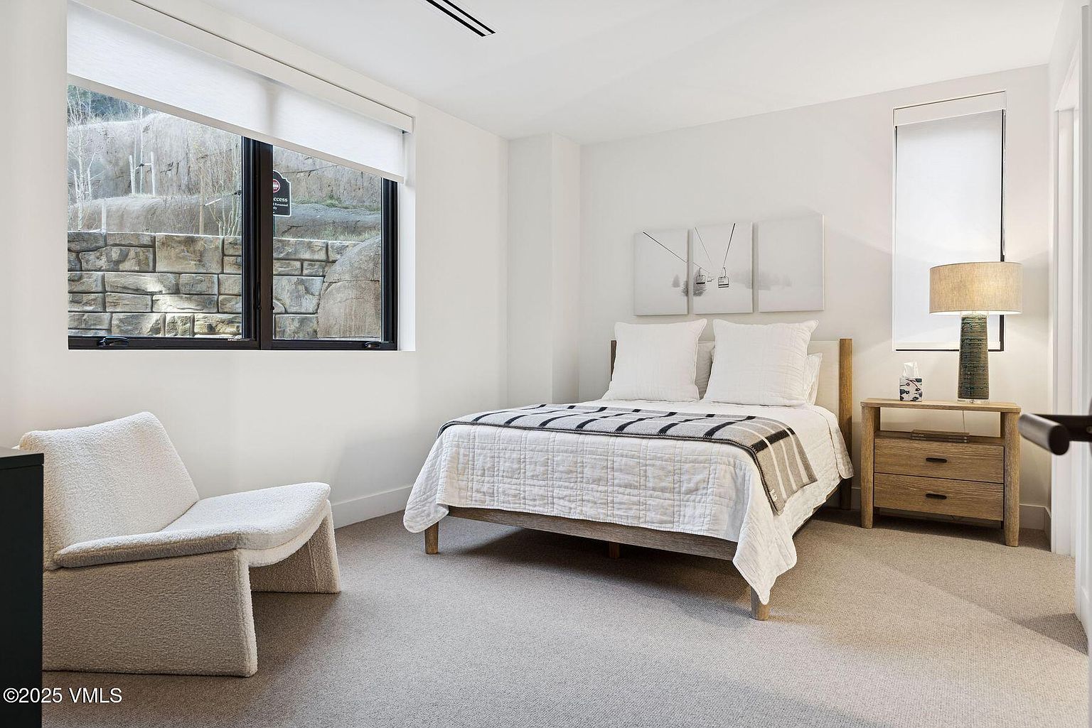 This is a bright and inviting guest bedroom featuring a neutral color palette. The room includes a bed with white linens and a patterned throw, a side table with a lamp, and a comfortable chair. A large window provides natural light and a view of the exterior, enhancing the room's serene atmosphere.