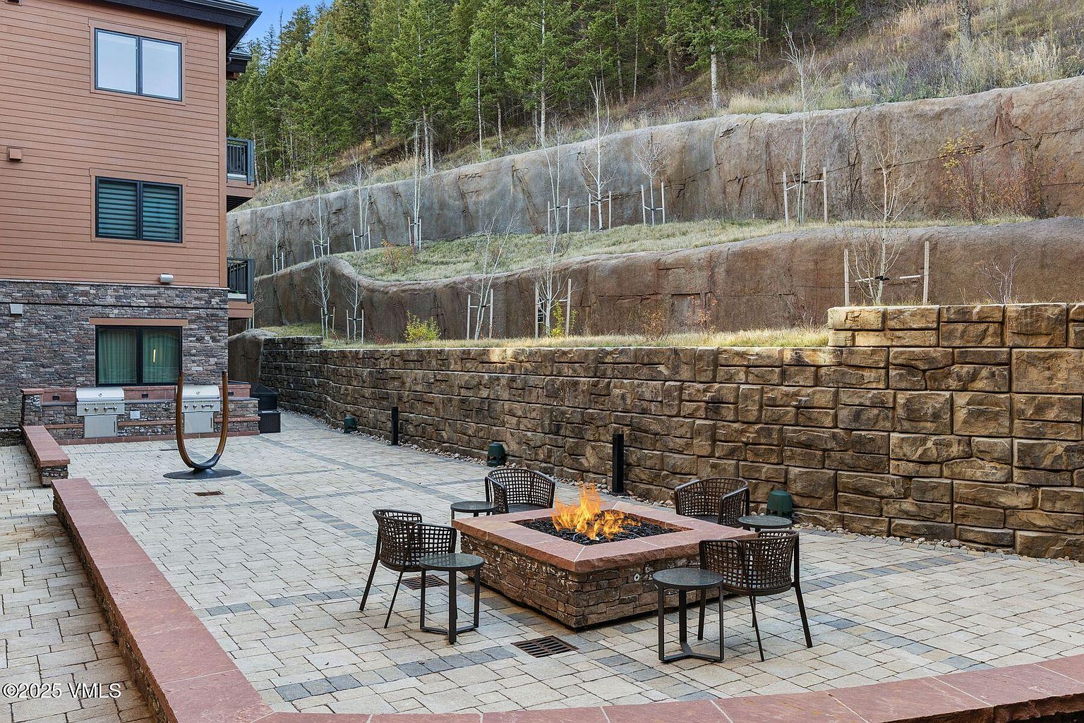 This image showcases a beautifully designed outdoor patio area, featuring a central fire pit surrounded by comfortable seating. The patio is constructed with patterned pavers and bordered by a low brick wall, creating a defined and inviting space. A stone retaining wall and lush greenery provide a scenic backdrop, enhancing the overall ambiance and privacy of the area.