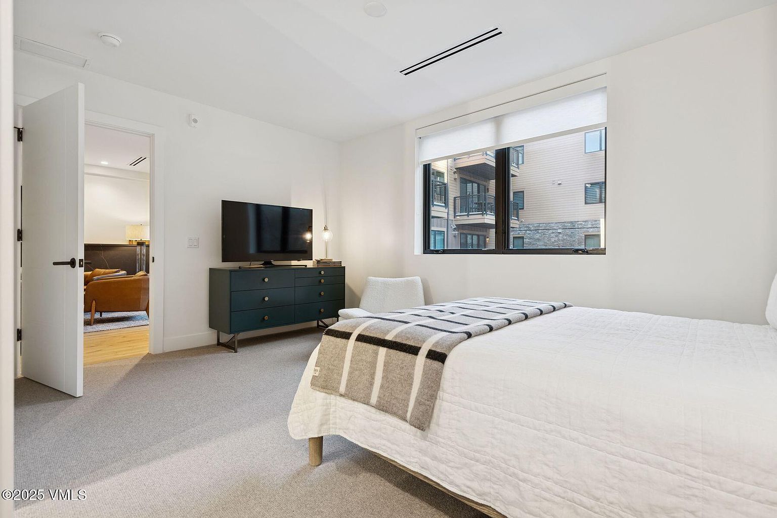 This is a bright and airy bedroom featuring a bed with a white quilt and a striped throw blanket. A dark green dresser with a television on top sits against the wall, next to a small chair. The room has a large window providing natural light and a view of the exterior buildings, and the walls are painted white, creating a clean and modern aesthetic.