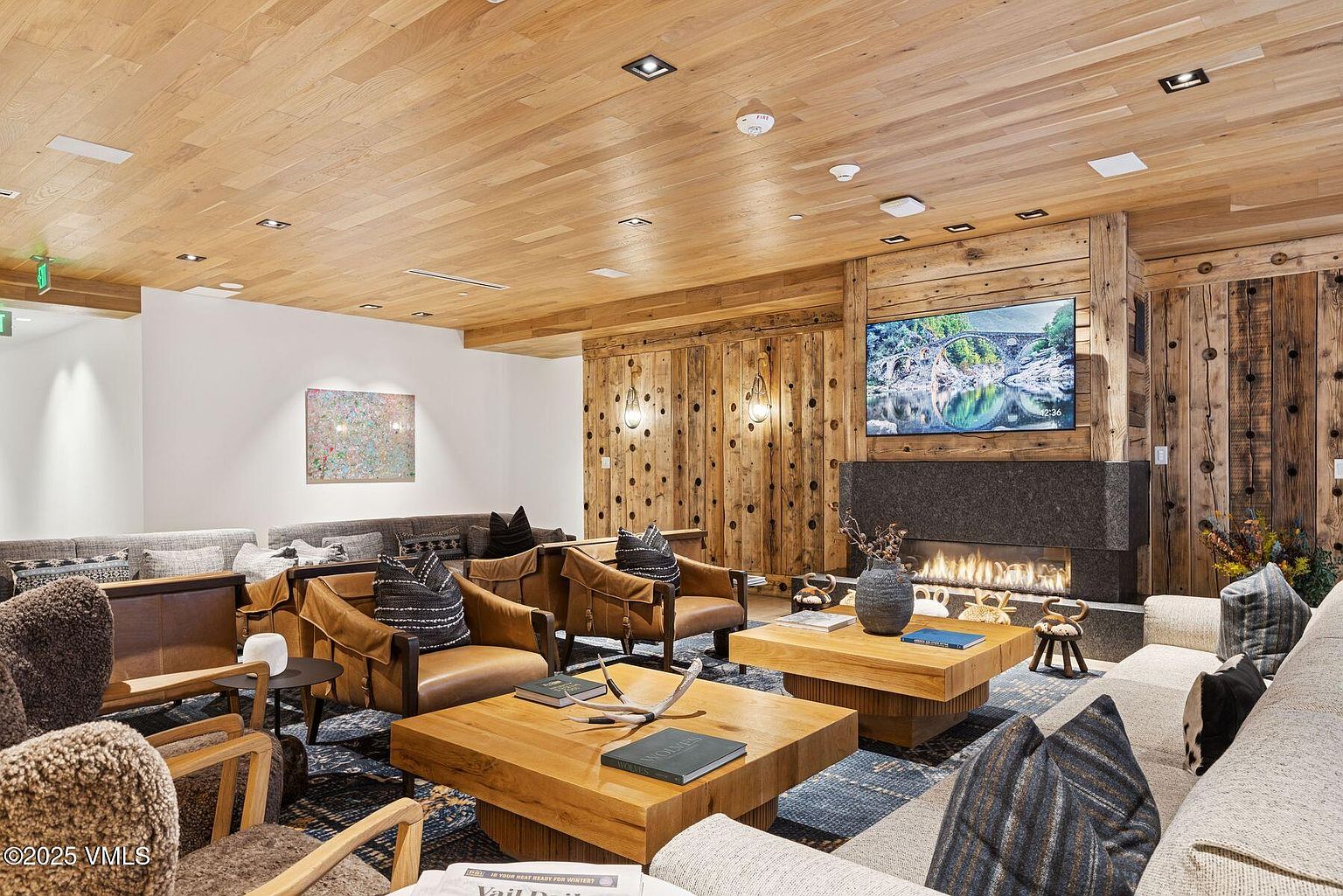 This inviting living room features a rustic yet modern design with wood-paneled walls and ceiling, complemented by comfortable leather armchairs and a large sectional sofa. A stone fireplace with a mounted television adds a focal point, while wooden coffee tables and a patterned rug enhance the cozy atmosphere. The room exudes warmth and sophistication, perfect for relaxation and entertaining.