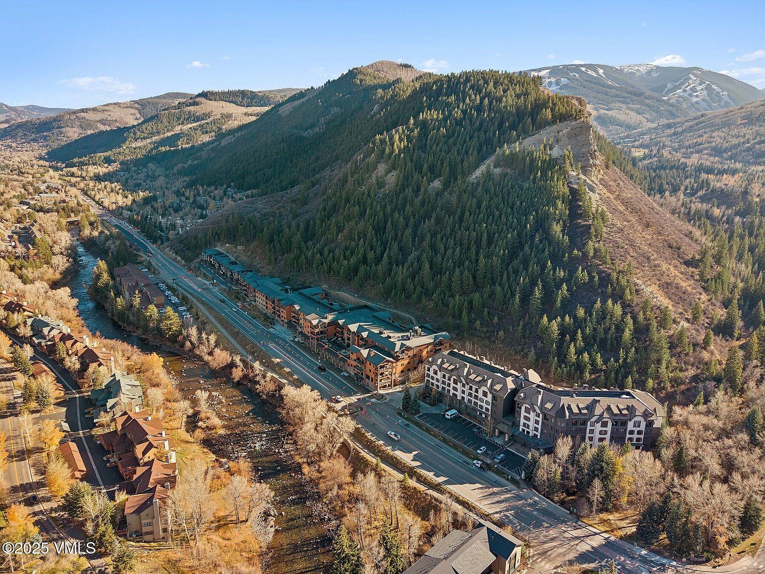 This aerial view showcases a luxurious mountain resort nestled in a valley, featuring multiple buildings with architectural details like gabled roofs and balconies. A winding river and a highway run alongside the resort, with lush green mountains providing a scenic backdrop. The overall impression is one of upscale living in a natural, serene environment.