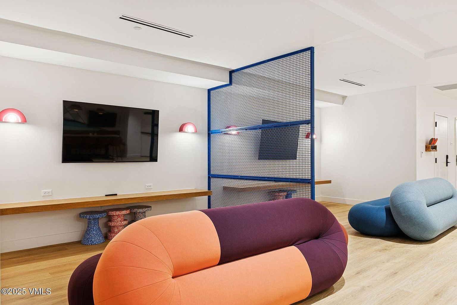 This interior shot showcases a modern living room with a playful design. The room features unique, colorful seating arrangements, a large flat-screen TV mounted on the wall, and a stylish blue metal grid room divider. The light wood flooring and minimalist decor contribute to a clean and inviting atmosphere.