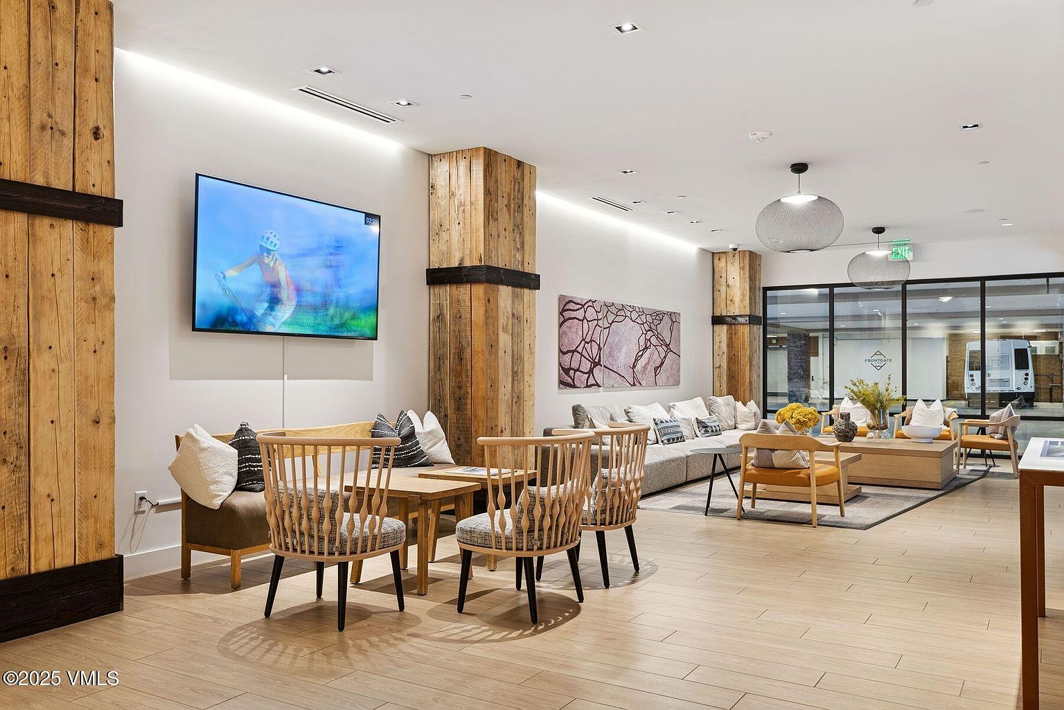 This is a well-lit and spacious living room area, featuring a blend of modern and rustic design elements. The room includes comfortable seating arrangements, a large television, and wooden support beams that add character. The flooring is light-colored wood, and the overall impression is inviting and stylish.