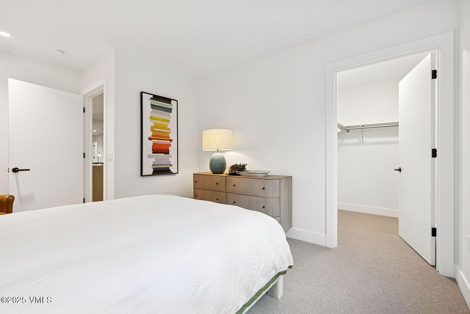 This is a bright and airy primary bedroom featuring a large bed with white linens and a green trim. A modern dresser with a lamp and artwork sits against the wall, and an open closet door reveals ample storage space. The room is painted in neutral tones, creating a clean and inviting atmosphere.