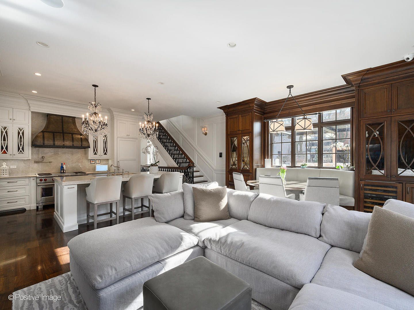 This expansive, open-concept living space seamlessly blends a modern kitchen, a dining nook, and a comfortable lounge area. The room features a large, plush grey sectional sofa, a sophisticated kitchen with white cabinetry and a dark range hood, and a dining area framed by rich, dark wood built-ins. The perspective captures the flow of the home, highlighting the elegant staircase and the bright, airy atmosphere created by the combination of natural light and refined architectural details.