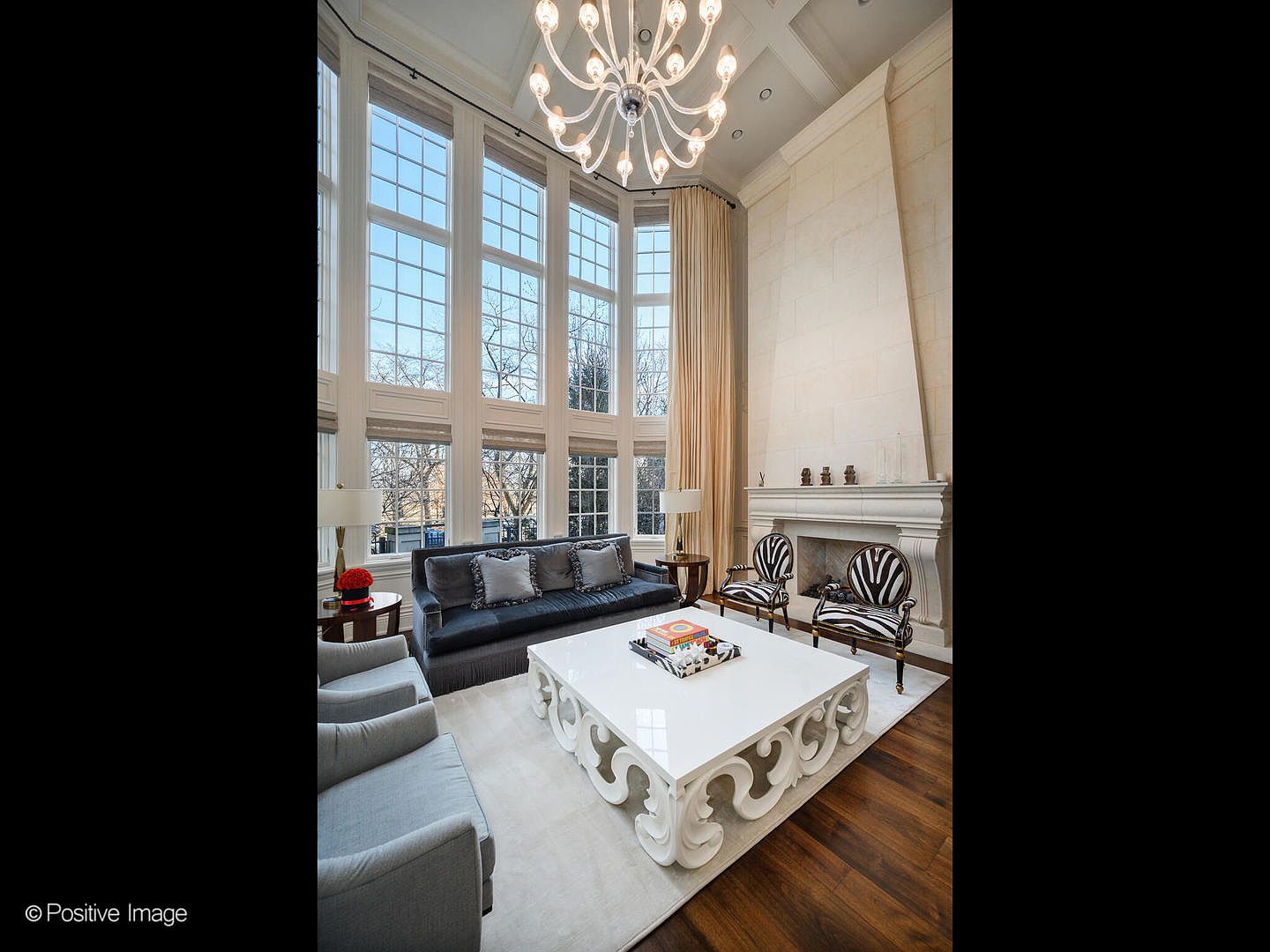 This grand, double-height living room features a dramatic floor-to-ceiling window wall that floods the space with natural light, complemented by a sophisticated crystal chandelier. The room is anchored by a large, ornate white coffee table and a sleek dark sofa, while a tall, limestone-style fireplace serves as a striking architectural focal point. The overall aesthetic is one of refined luxury, blending classic elegance with modern, high-contrast design elements.