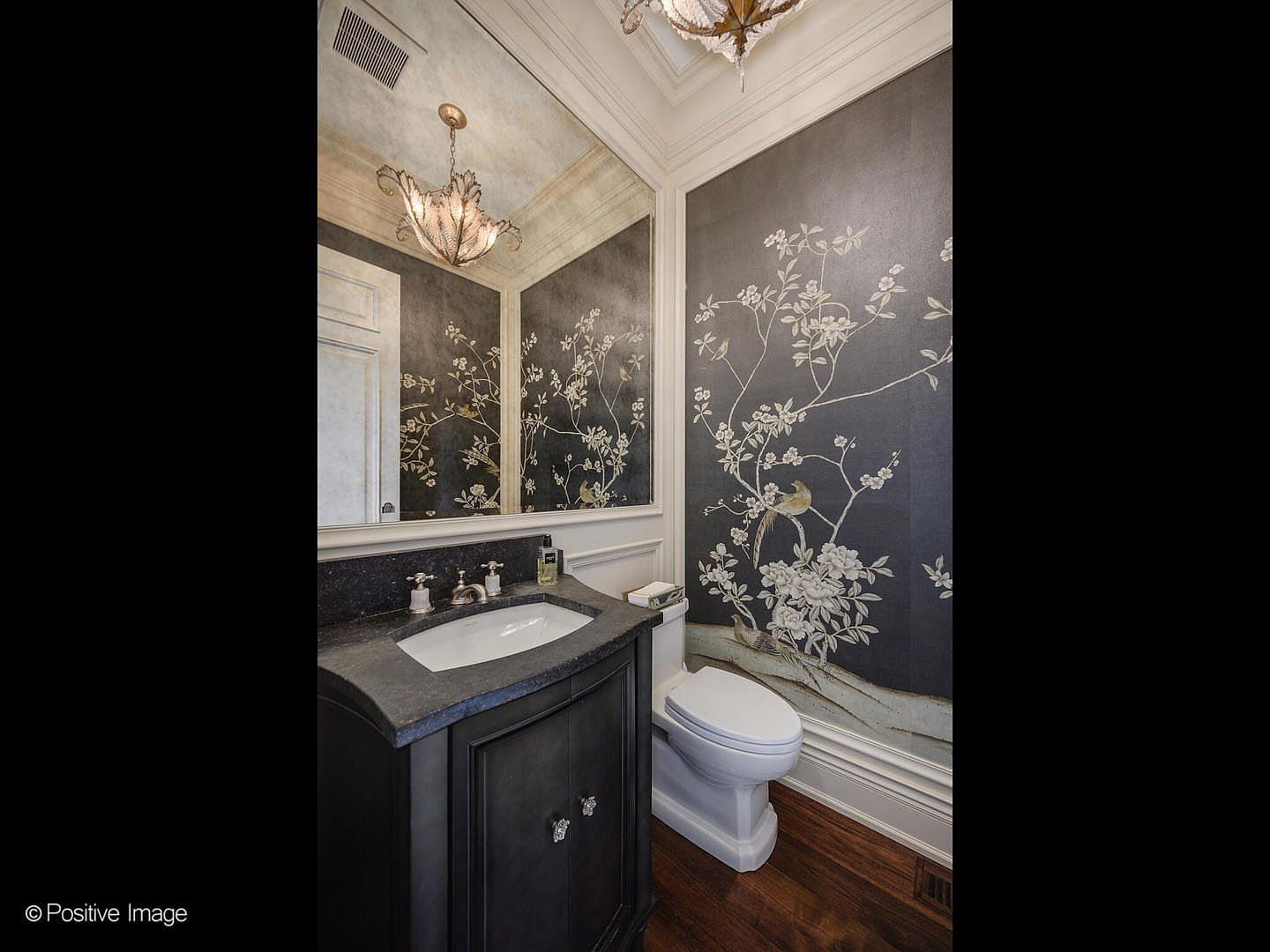 This elegant powder room features sophisticated dark floral wallpaper that creates a dramatic and luxurious atmosphere. The space is anchored by a dark wood vanity with a stone countertop and a classic white undermount sink, complemented by a large mirror and a decorative chandelier. The combination of the ornate wall covering and traditional fixtures gives the room a refined, high-end aesthetic.