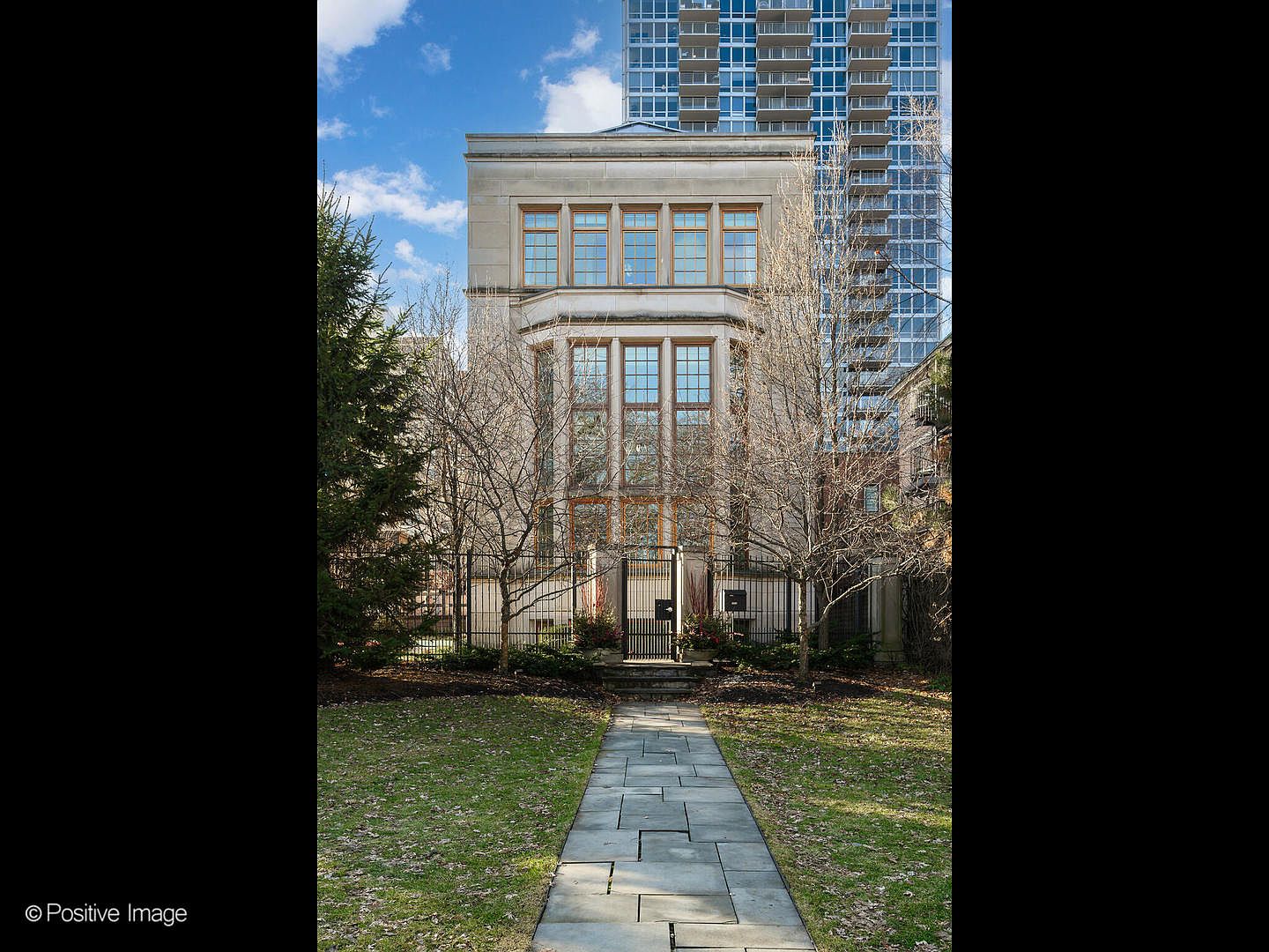 This image captures a stately, multi-story stone residence featuring a symmetrical facade with large, multi-pane windows and a prominent central bay. A stone pathway leads directly to a wrought-iron gate, framed by mature trees and a manicured lawn, creating a sense of privacy and elegance. The composition is balanced and formal, with a towering modern high-rise visible in the background, highlighting the property's unique urban setting.