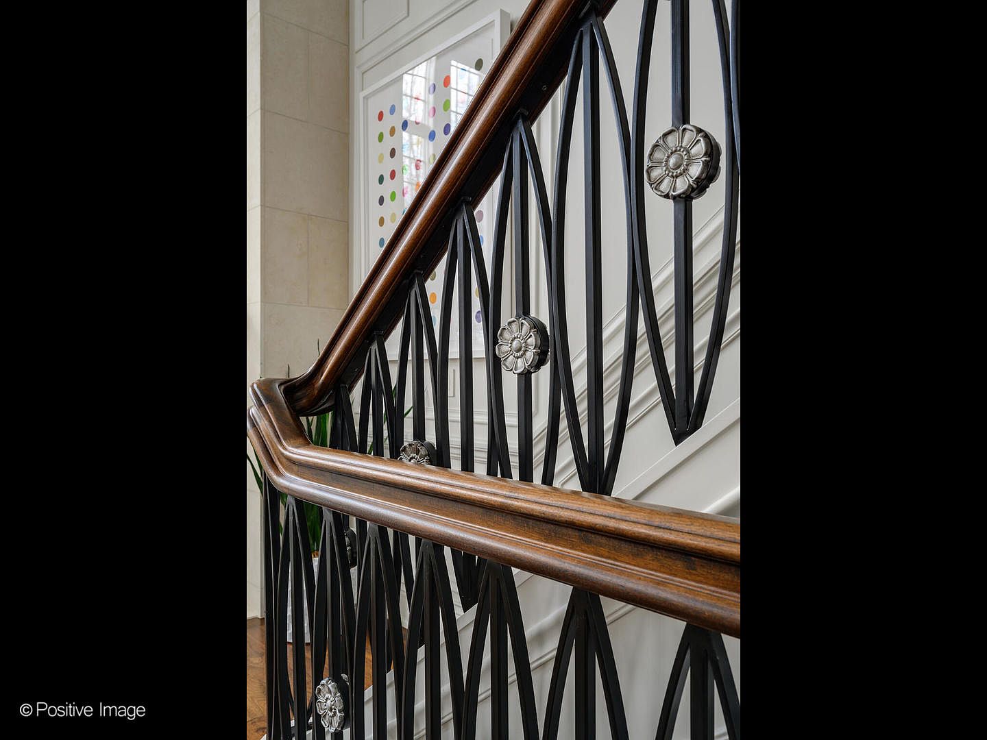 This close-up shot highlights an elegant staircase railing featuring dark, intricate metalwork accented with decorative silver floral medallions. The rich, polished wooden handrail curves gracefully, leading the eye upward toward a bright, art-filled wall in the background. The composition emphasizes high-end craftsmanship and sophisticated interior design details.