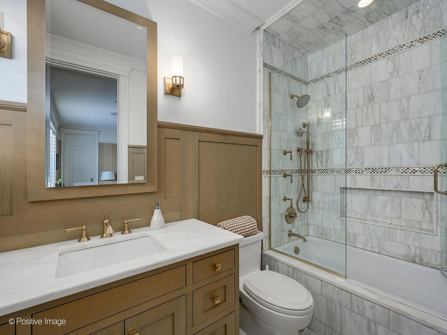 This elegant bathroom features a custom wood vanity with a marble countertop and brass fixtures, complemented by wainscoting on the walls. A glass-enclosed shower and tub combination is tiled in white marble with a decorative mosaic accent strip, creating a sophisticated and cohesive aesthetic. The perspective is a medium-wide shot capturing the vanity, toilet, and shower area, emphasizing the high-end finishes and clean design.