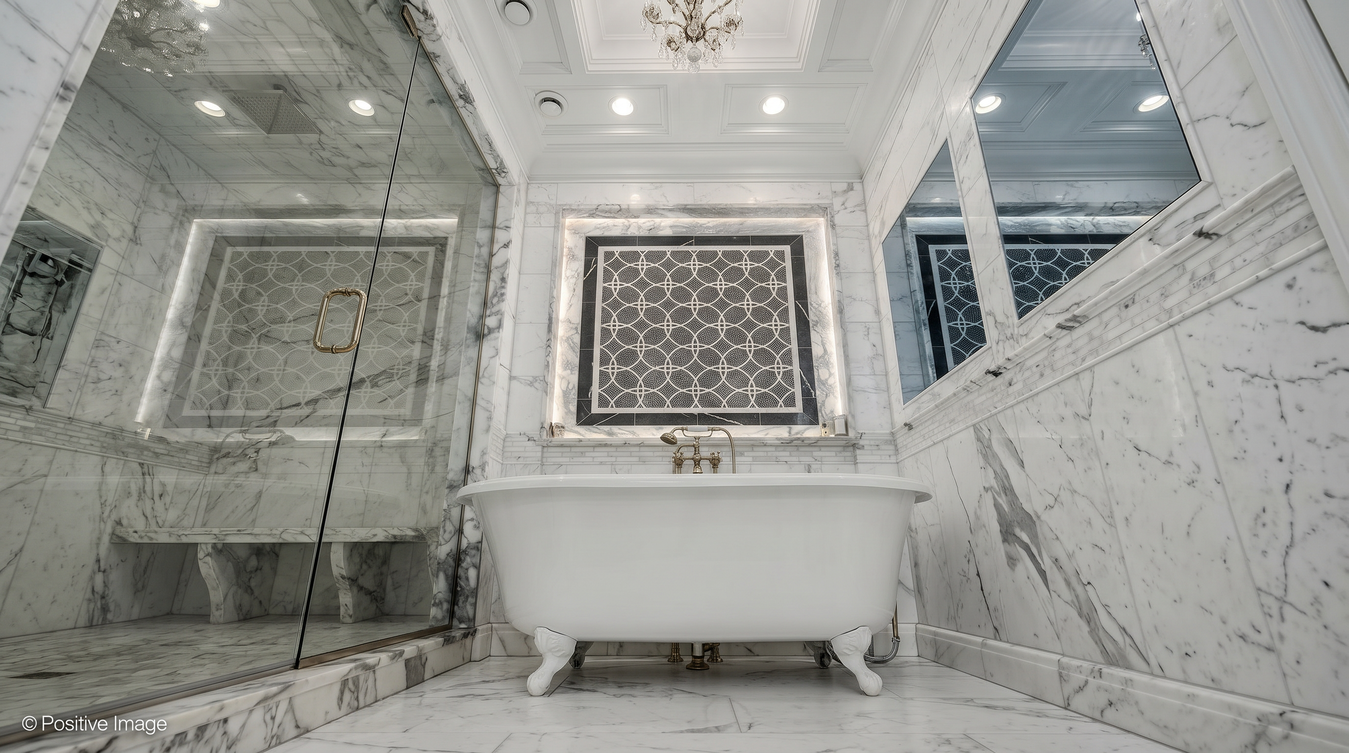 This luxurious primary bathroom is defined by its extensive use of high-end white marble with dramatic grey veining. The focal point is a classic clawfoot soaking tub centered beneath an intricately patterned mosaic wall inlay, complemented by a spacious glass-enclosed shower and elegant gold-toned fixtures. The room radiates opulence with its coffered ceiling, recessed lighting, and a crystal chandelier, creating a refined and high-end spa-like atmosphere.