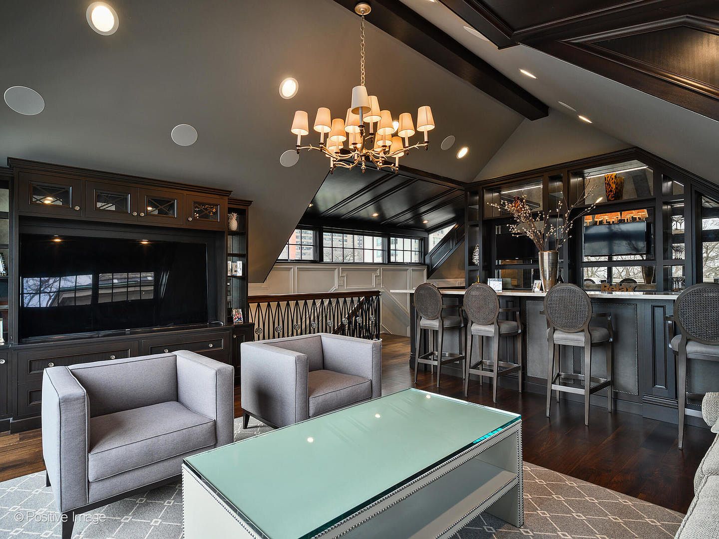 This sophisticated living room and bar area features a high, vaulted ceiling with dark wood beams and a grand chandelier, creating an elegant and moody atmosphere. The space is anchored by a large built-in entertainment center, two plush grey armchairs, and a modern glass-topped coffee table, while a sleek bar with four stools provides a perfect spot for entertaining. The dark cabinetry and rich hardwood floors contrast beautifully with the light grey furniture, offering a luxurious and well-appointed aesthetic.