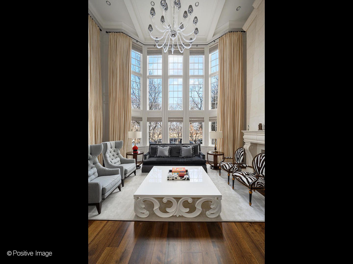 This grand, double-height living room exudes luxury with its soaring floor-to-ceiling windows draped in elegant floor-length curtains and a dramatic, intricate crystal chandelier. The space is anchored by a large, glossy white coffee table, flanked by a plush grey sofa and a mix of contemporary grey armchairs and striking zebra-print accent chairs. The room features rich hardwood flooring and a classic stone fireplace, creating a sophisticated and airy atmosphere perfect for formal entertaining.