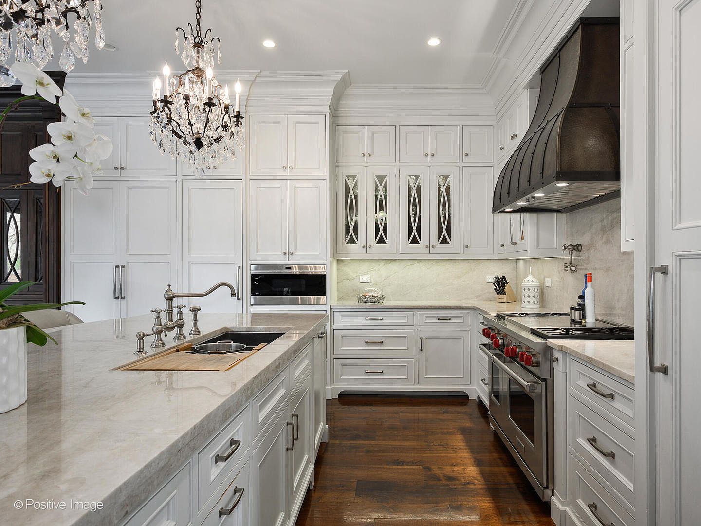 This luxurious, high-end kitchen features crisp white cabinetry, a large marble-topped island, and a striking dark metal range hood that serves as a focal point. The space is elegantly appointed with a crystal chandelier, professional-grade stainless steel appliances, and warm hardwood flooring, creating a sophisticated and inviting atmosphere. The perspective is a wide-angle shot from the kitchen island, showcasing the seamless flow between the prep area and the main cooking station.