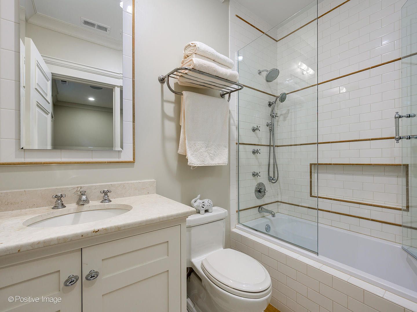 This bright and clean bathroom features a classic vanity with a marble countertop, a white toilet, and a shower-tub combination enclosed by a glass panel. The walls are adorned with white subway tiles accented by thin gold trim, creating a sophisticated and timeless aesthetic. The perspective is a straight-on shot that captures the functional layout and elegant finishes of the space.