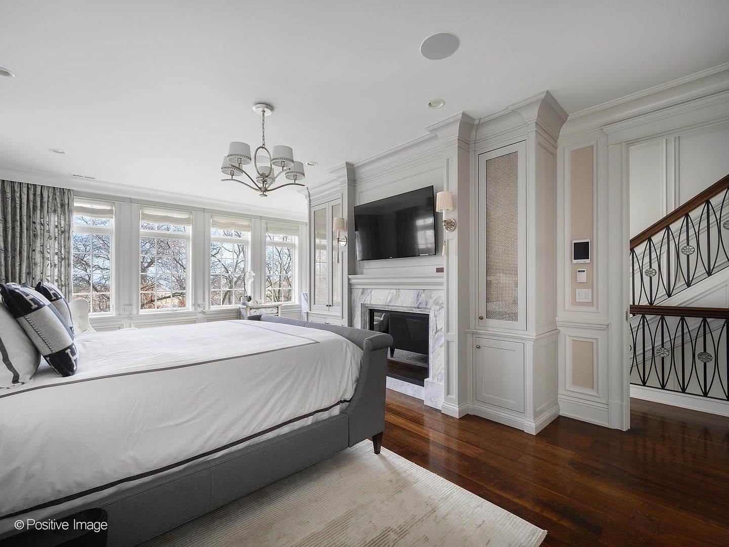 This elegant primary bedroom features a sophisticated gray upholstered bed, a marble-surround fireplace with an integrated television, and custom built-in cabinetry. Large, multi-pane windows allow for ample natural light, while the rich dark hardwood floors and classic architectural molding create a refined, luxurious atmosphere. The perspective is captured from the foot of the bed, looking toward the fireplace and the adjacent staircase, emphasizing the room's spacious and well-appointed layout.