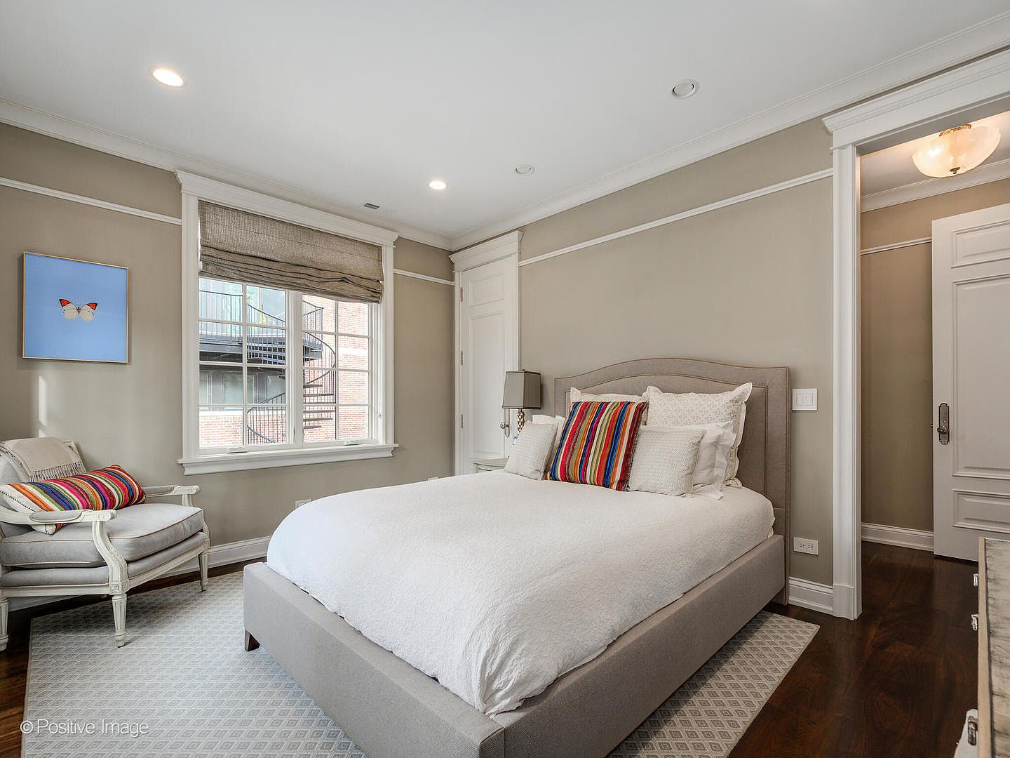 This inviting bedroom features a neutral color palette with a plush, upholstered bed as the focal point, accented by vibrant, multi-colored striped pillows. A cozy armchair sits near the window, which is dressed in a textured Roman shade, while dark hardwood floors provide a rich contrast to the light walls. The room is well-lit with recessed lighting and a classic bedside lamp, creating a warm and comfortable atmosphere.