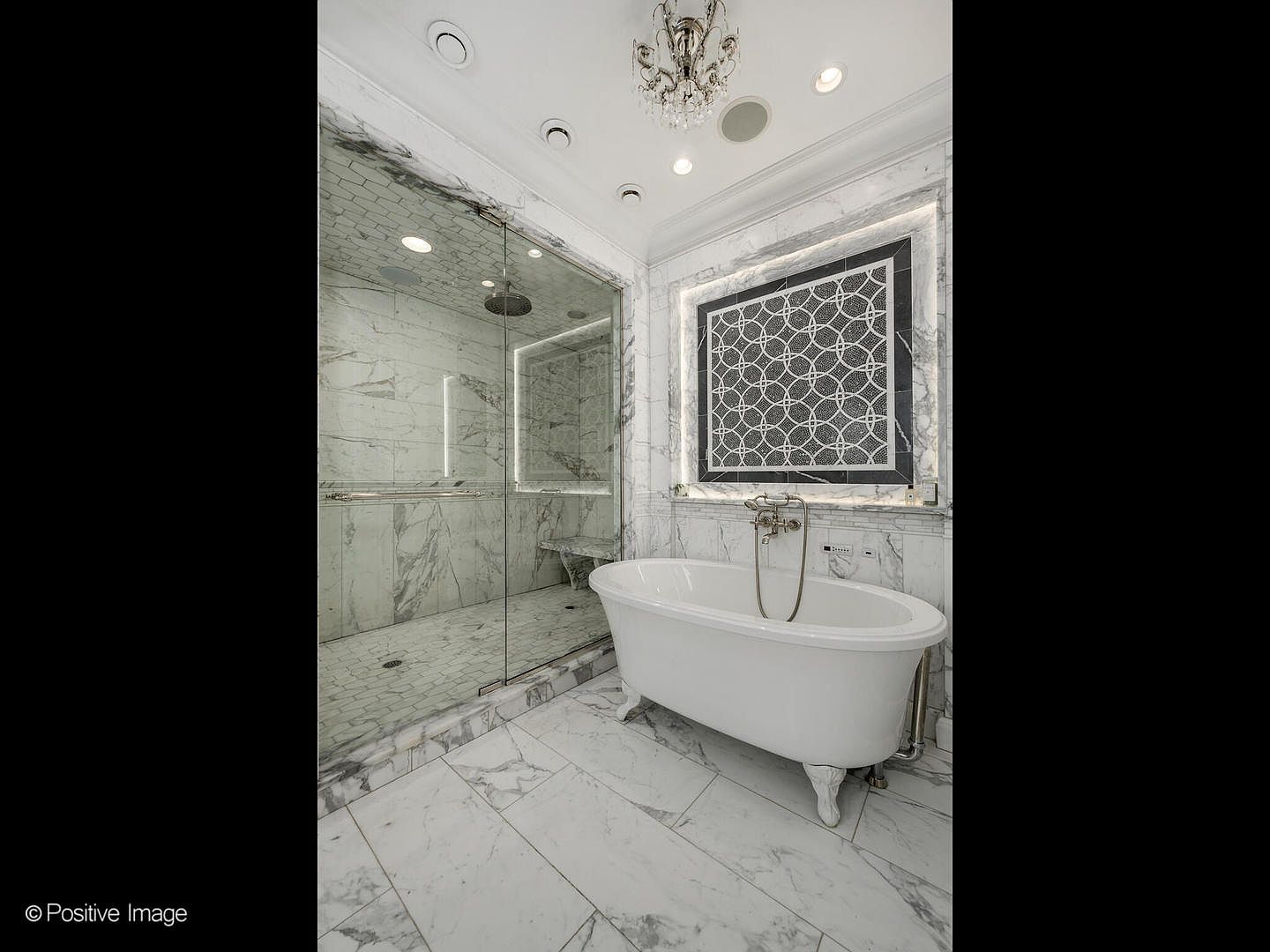 This luxurious primary bathroom features a sophisticated design with floor-to-ceiling marble tiling and a classic clawfoot soaking tub as the centerpiece. To the left, a spacious walk-in glass shower is integrated into the layout, while an elegant geometric mosaic tile accent wall behind the tub adds a touch of modern artistry. The room is illuminated by recessed lighting and a delicate crystal chandelier, creating an atmosphere of refined elegance and spa-like tranquility.