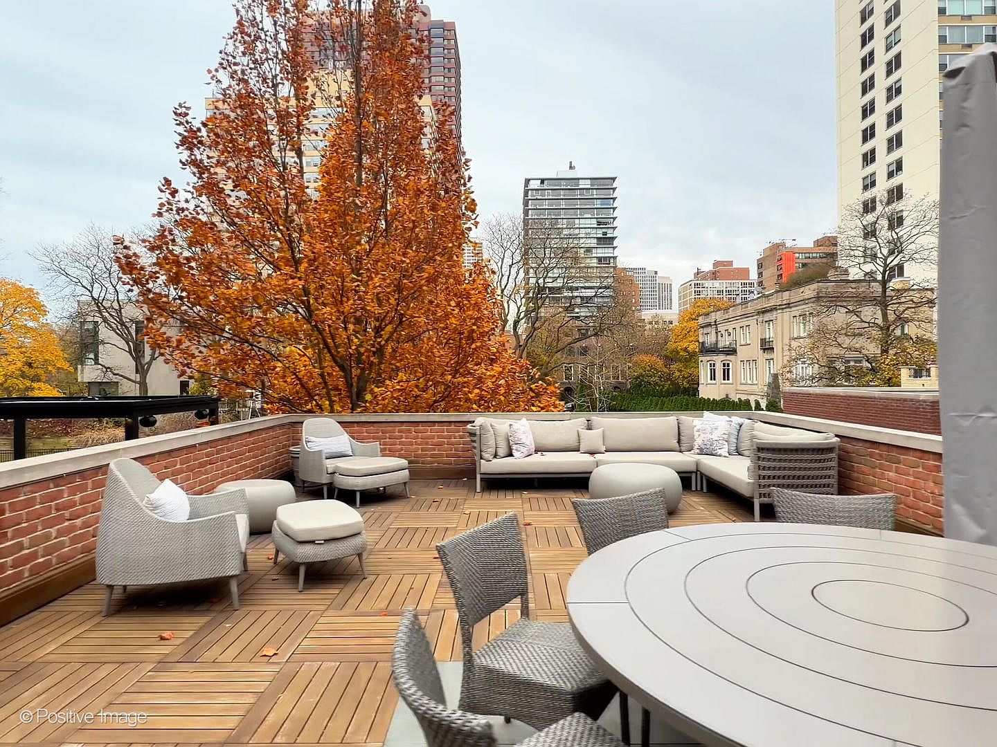 This spacious rooftop terrace features high-end, modern outdoor furniture, including a large sectional sofa, armchairs with ottomans, and a circular dining table. The deck is finished with warm-toned wooden decking and is enclosed by a classic brick parapet wall. The setting offers a sophisticated urban retreat with sweeping views of surrounding high-rise buildings and vibrant autumn foliage.