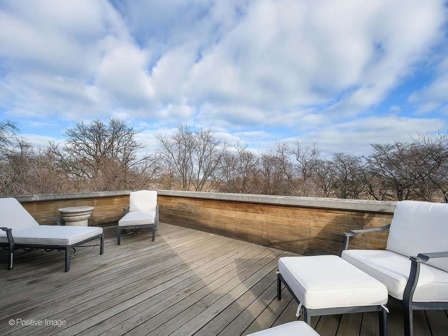 This spacious rooftop deck features weathered wood decking and a clean, minimalist aesthetic, furnished with comfortable white lounge chairs and an ottoman. The area is enclosed by a low wooden wall with a stone cap, offering an unobstructed, elevated view of a dense, leafless forest under a bright, cloudy sky. The perspective is wide and inviting, capturing the serene and private atmosphere of this outdoor living space.
