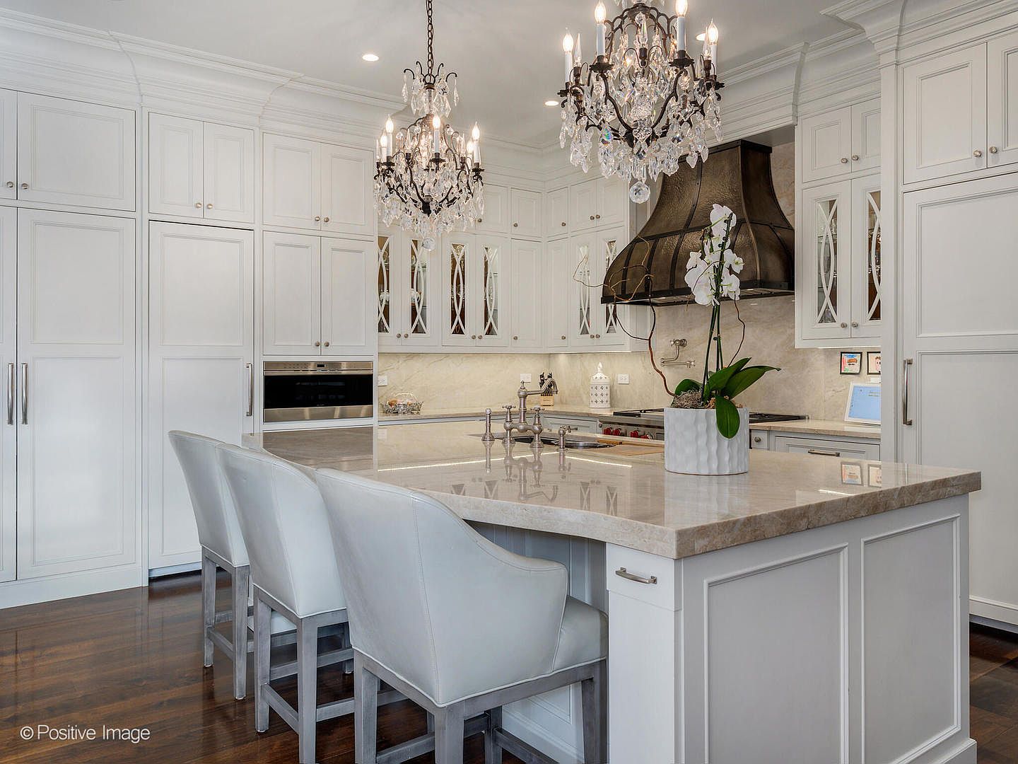 This elegant, high-end kitchen features crisp white cabinetry, a large stone-topped island with comfortable bar seating, and a striking dark metal range hood. Two ornate crystal chandeliers hang above the island, adding a touch of luxury to the bright, sophisticated space. The perspective is a medium shot from the island, showcasing the seamless blend of classic design and modern functionality.
