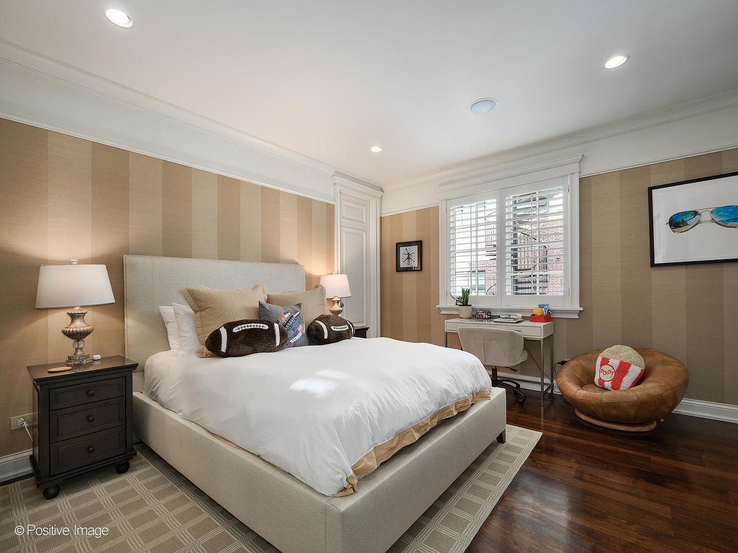 This well-appointed bedroom features a neutral color palette with striped wallpaper, a plush upholstered bed, and dark hardwood floors. The room includes a dedicated workspace with a desk and chair, as well as a cozy leather lounge chair, creating a versatile and inviting atmosphere. The perspective is a wide-angle shot from the foot of the bed, capturing the entire room's layout and natural light streaming through the window shutters.