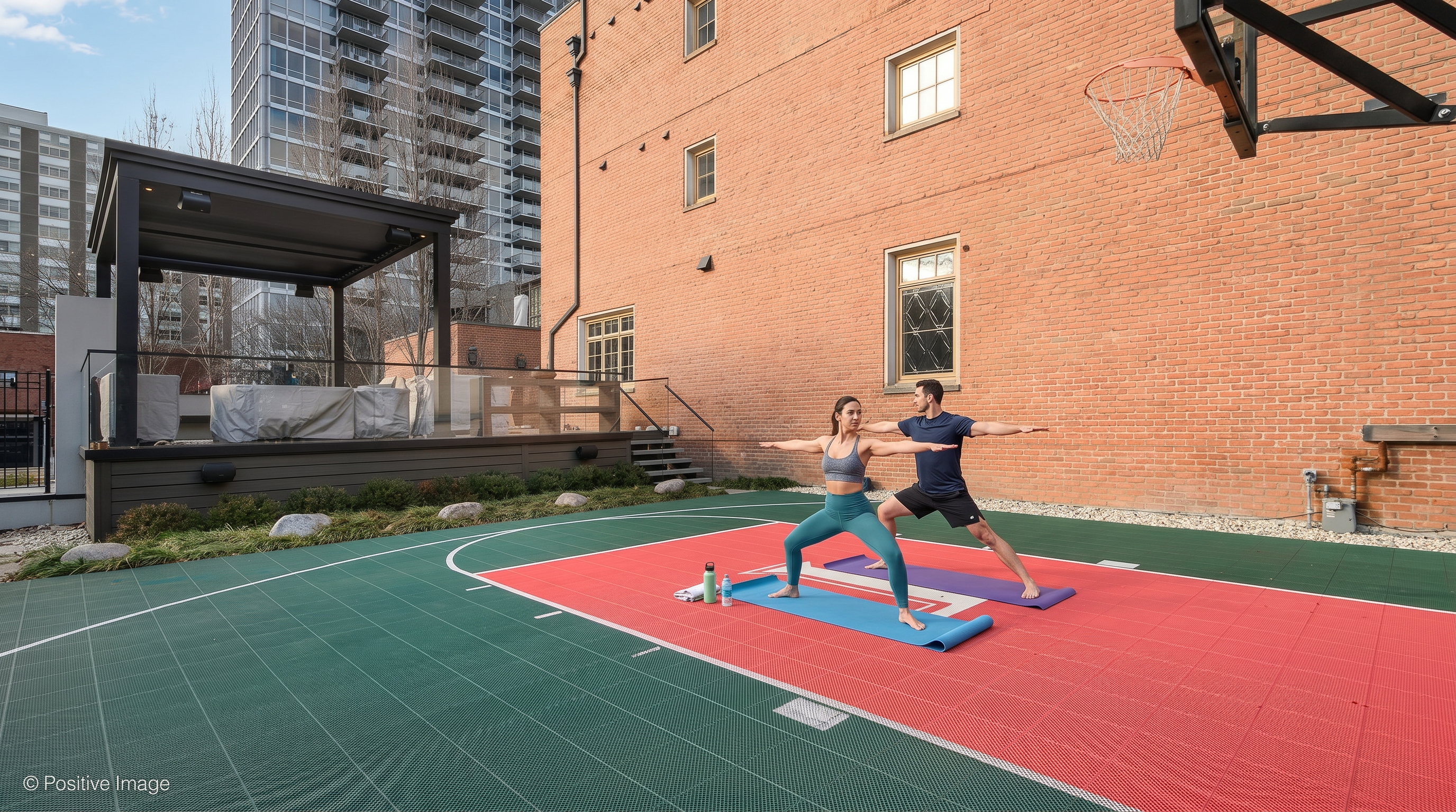 This outdoor community amenity space features a paved, multi-use recreation area, identified by sports court markings, where two individuals are performing yoga. Adjacent to the recreational area is a covered patio space with contemporary styling and comfortable outdoor furniture. The scene, framed by historic brick architecture and modern high-rise buildings, illustrates an engaging environment for resident fitness and social activity.