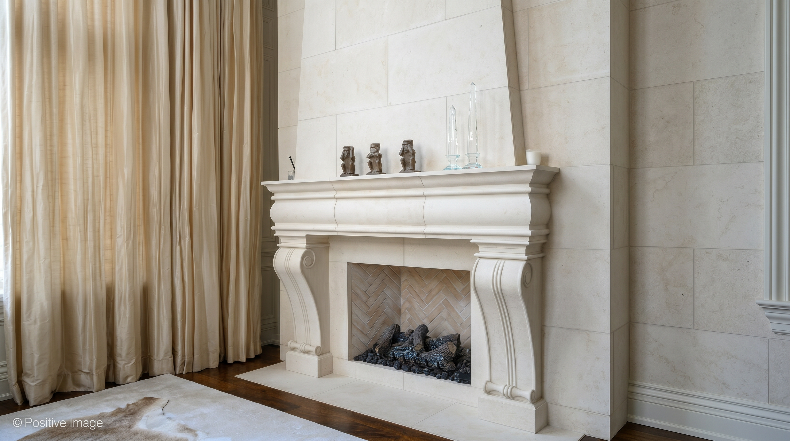 This image highlights a grand, custom-carved stone fireplace mantel that serves as a sophisticated focal point for the room. Featuring elegant curved corbels and a classic, neutral aesthetic, it is set against a wall of matching tiled stone with a herringbone-patterned inner firebox. The mantle is tastefully decorated with subtle ornamental figurines and glass obelisks, exuding an air of luxury and timeless refinement.