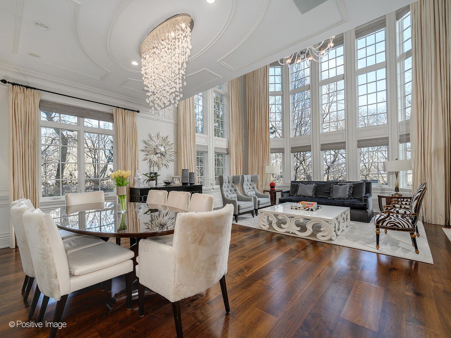 This expansive, high-ceilinged living and dining area exudes luxury with its grand crystal chandelier, floor-to-ceiling windows, and rich hardwood flooring. The space features a sophisticated blend of formal dining furniture and a plush, contemporary seating area anchored by a large, ornate coffee table. The bright, airy atmosphere is enhanced by elegant drapery and a neutral color palette, creating a refined and welcoming environment.