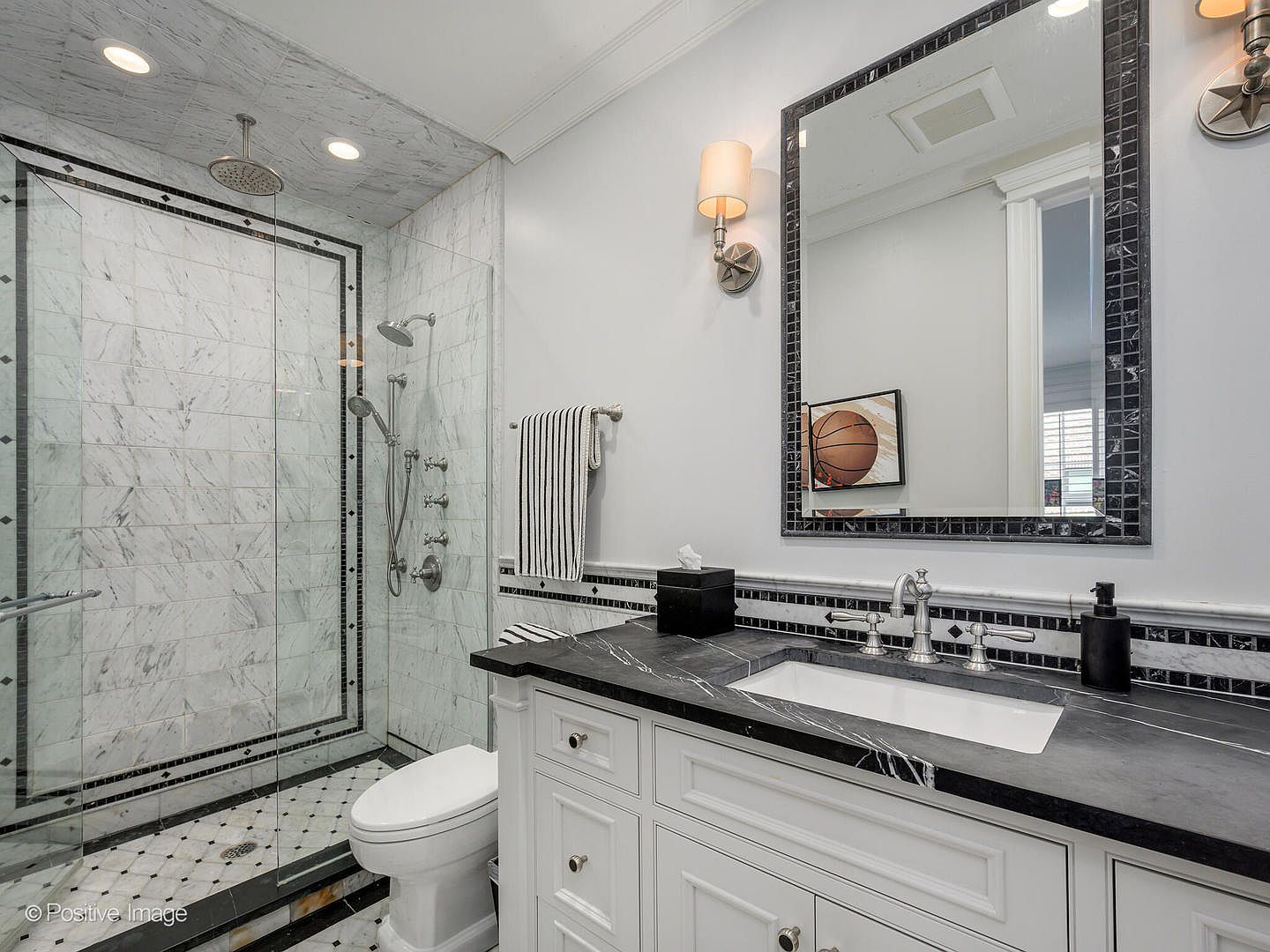 This elegant bathroom features a sophisticated black and white color palette, highlighted by a marble-tiled walk-in shower with a rainfall showerhead and a sleek black stone vanity countertop. The space is accented with classic white cabinetry, a framed mirror, and decorative wall sconces, creating a polished and timeless aesthetic. The perspective is a wide-angle shot from the doorway, capturing the seamless integration of the shower, vanity, and toilet area.