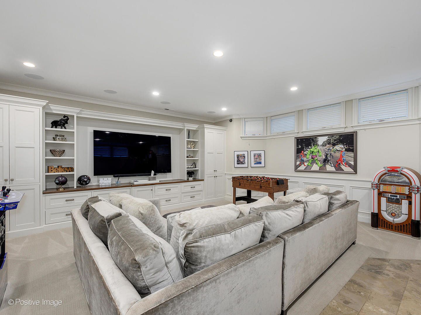 This spacious, well-lit basement recreation room features a large, plush sectional sofa facing a built-in media center with a flat-screen television. The room is designed for entertainment, complete with a foosball table, a vintage-style jukebox, and a game console, all set against a backdrop of custom white cabinetry and framed superhero artwork. The neutral color palette and recessed lighting create a comfortable, inviting atmosphere perfect for family gatherings or hosting guests.